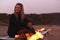 Woman Sitting On Surfboard By Camp Fire On Beach As Sun Sets Behind Her