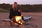 Woman Sitting On Surfboard By Camp Fire On Beach Using Mobile Phone As Sun Sets Behind Her