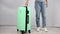 The woman spins a mint plastic suitcase on wheels and leaves.