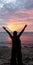 A woman standing on the beach with her hands raised in silhouette, enjoying the sunset sky in the distance at dusk.