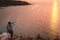 A woman is standing on the edge of a cliff and taking photos of the sunset on her smartphone. In the background, the sea at sunset