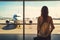 Woman standing at lounge with luggage watching at airport window while waiting at boarding gate before departure. AI