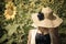 Woman in straw hat with big blue faux flower with head down in field near big sunflower flower.