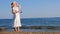 woman in a straw hat and white dress takes off her sunglasses and enjoys the sun while walking along the seashore and