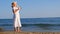 woman in a straw hat and white dress walks along the seashore puts on sunglasses and admires the blue sea on a sunny