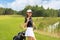 Woman in stylish summer golf outfit walking with bag of drivers on beautiful green course for golf game.