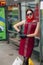Woman in sunglasses, in a red dress, stands with groceries in bags, at the bus station.