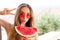 Woman in a swimsuit and red sunglasses with a watermelon shows a peace sign on a background of tropical hotels and the beach