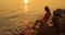 Woman in swimsuit sitting on cobblestone and wetting her feet in sea at sunset.