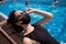 Woman in swimsuit, sunglasses and protective black mask sunbathes against backdrop of hotel pool on sun lounger