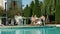 Woman taking video call by the pool and group of teens talking at another table while young woman relaxes on the pool edge