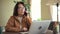 Woman talking on phone and working with laptop at table in home office indoors.