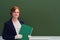 Woman teacher holds a green folder while standing near the school blackboard, copy space