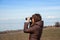 Woman tourist looking through binoculars at distant sea, enjoying landscape. Autumnal time.Lonely woman in brown coat