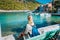 Woman tourist sitting on the bank in front of beautiful sea bay on summer morning. Lovely vacation in Assos village with