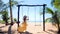 Woman tourist swinging on swing on sandy beach with palm trees and blue sea