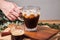 woman used hand take cold iced black brew coffee in a glass with pieces of ice on a wooden table, natural light, fresh summer