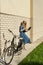 Woman and vintage bicycle and white brick wall, green lawn. Blonde girl is standing outside an office, holding a tablet