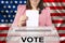 Woman voter in smart pink jacket lowers the ballot in a transparent ballot box against the background of the national flag of the