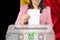Woman voter in a smart pink jacket lowers the ballot in a transparent ballot box against the background of the national flag of