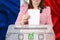 Woman voter in a smart pink jacket lowers the ballot in a transparent ballot box against the background of the national flag of
