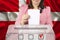 Woman voter in smart pink jacket lowers the ballot in a transparent ballot box against the background of the national flag of the