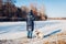 Woman walking pug dog in snowy winter park by frozen lake holding leash. Puppy wearing harness