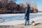 Woman walking pug dog in snowy winter park by frozen lake holding leash. Puppy wearing harness