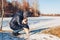 Woman walking pug dog in snowy winter park by frozen lake hugging pet. Happy puppy wearing harness