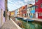 Woman walks in Burano island picturesque street with small colored houses in row, water canal with fishermans boats, c