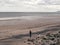 Woman walks down a beach looking at her phone in her hand with a dog. Cold with