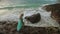 Woman walks on rock of sea reef stone, stormy cloudy ocean. Blue swimsuit dress tunic. Concept rest, tropical resort