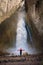 A woman in a warm jacket and hat stands with her back against the background of a huge epic waterfall flowing from large