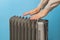 Woman warms her hands near hot oil filled electric heater against blue background. Using 9 fins space heater. Portable household