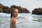 Woman with waterproof makeup submerges in ocean, stones backdrop. Female tours seascape, enduring swimwear stylish
