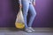 woman wearing casual clothes holding string shopper bag with fresh bananas against violet wall, eco friendly and healthy