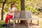 Woman Wearing Fitness Clothing Sitting On Seat Under Tree Checking Activity Monitor On Smartwatch