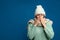 Woman wearing hat and scarf sneezing on blue background, space for text. Cold symptoms