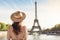 A woman wearing a hat stands in front of the iconic Eiffel Tower, tourist woman in summer dress and hat standing on beautiful