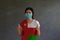 Woman wearing hygienic mask and wearing Oman flag colored shirt with thumbs up with both hands on dark wall background