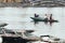 Woman wearing purple long arm t-shirt and conical hat rowing boat by her feet with tourists on the boat on the river at Trang An.