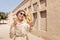 Woman wearing a turban hat takes a selfie against the backdrop of a desert fortress city on the Arabian Peninsula