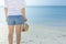 Woman wearing white t-shirt, she standing on sand beach and holding weave hat in hand, she looking at the sea and blue sky.