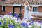 Woman in white dress holding basin near ancient artesian draw-well in village. Colorful violet flowers on foreground. Girl gets