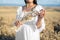 Woman in white dress stands in field with wheat. Person hold in hands bundle of ripe spikelets. Harvest season.