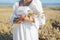 Woman in white dress stands in field with wheat. Person hold in hands bundle of ripe spikelets. Harvest season.
