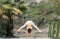 A woman in a white gymnastic jumpsuit is engaged in yoga on the background of water and a green mountain slope