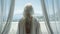 Woman with white hair looking out from a bright window with sea view.