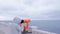 Woman in white jacket and hood looks in binoculars at sea waterfront with sea view. Side view.