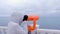 Woman in white jacket and hood looks in binoculars at sea waterfront with sea view. Side view.